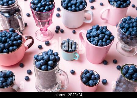 Bleuets biologiques en tasses et verres de différentes tailles et couleurs. Fruits d'été fraîchement récoltés. Une abondance de bleuets dans des boîtes sur un rose Banque D'Images