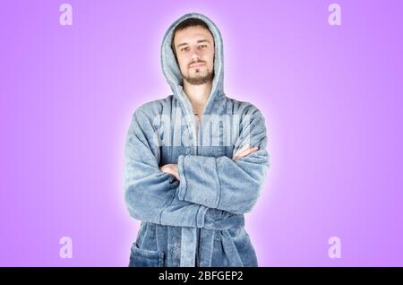 Portrait de jeune homme barbu caucasien dans un peignoir bleu avec des mains croisées isolées sur fond violet. Concept d'émotion confiant Banque D'Images