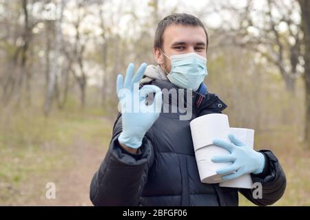 Concept Covidiot. Jeune homme dans le masque de protection tient beaucoup de rouleaux de papier toilette et montre bon geste à l'extérieur dans le bois de printemps. Achat de panique pendant qu Banque D'Images