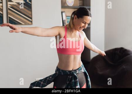 Jeune femme active dans des vêtements de sport dansant dans le salon avec intérieur confortable et moderne, profitez du concept de vie. Occupation pendant l'isolement à la maison. Banque D'Images