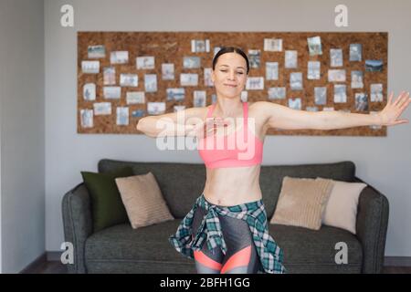 Jeune femme active dans des vêtements de sport dansant dans le salon avec intérieur confortable et moderne, profitez du concept de vie. Occupation pendant l'isolement à la maison. Banque D'Images