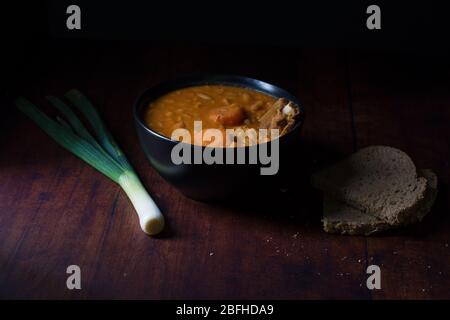 Côtes de porc fumées cornées avec haricots et morceaux de carottes, dans un bol noir, avec deux morceaux de pain de grains entiers couchés à côté, et un oignon vert Banque D'Images