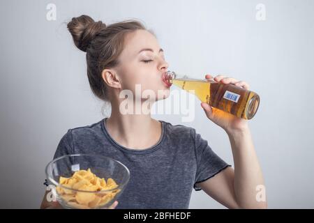 Jeune femme caucasienne souriante qui boit une bouteille de bière et qui contient des chips de pommes de terre Banque D'Images