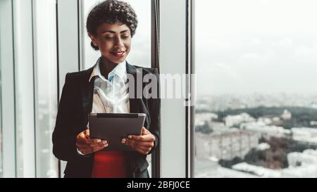 Une femme d'affaires comely afro-américaine avec une tablette numérique entre ses mains se penche contre le faisceau de fenêtre avec un horizon urbain à l'extérieur Banque D'Images