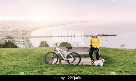 Femme asiatique en montée avec VTT. Une femme utilisant un téléphone cellulaire à l'affût d'un chien à Oamaru, Nouvelle-Zélande. Banque D'Images