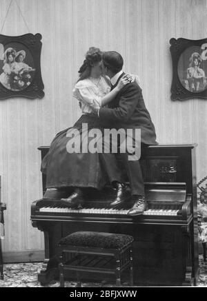 Couple baiser et embrasser, tout en étant assis sur un piano droit, les pieds du couple reposant sur le clavier. 1907. Pour voir toutes mes images liées à l'amour - dans la recherche: Prestor vintage love music Banque D'Images