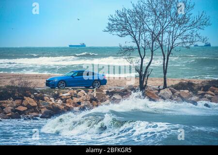 Vagues qui s'écrasent sur la côte. Homme conduisant voiture de sport se détendre près de la plage. Banque D'Images