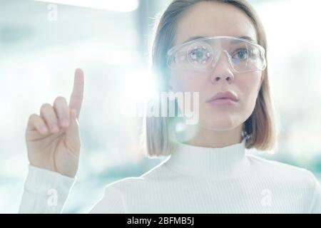 Jolie jeune femme développeur de technologie dans des lunettes intelligentes en appuyant sur le bouton virtuel à l'écran tout en faisant la présentation Banque D'Images