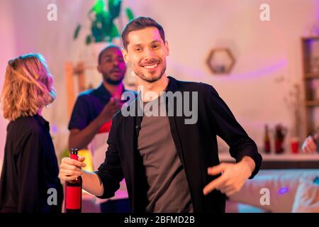 Jeune homme gai avec un sourire taothy tenant une bouteille de bière tout en dansant devant la caméra sur fond d'amis interculturels Banque D'Images