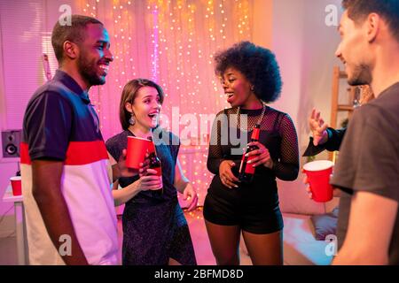 Deux jeunes couples interculturels joyeux dans une tenue glamour avec des boissons à la fête dans l'environnement domestique et discuter de choses drôles Banque D'Images