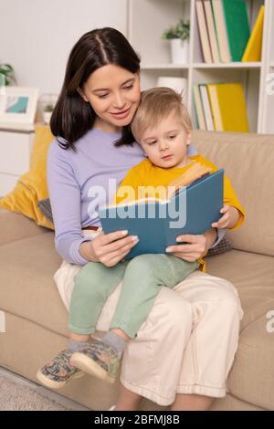 Heureuse jeune femme soigneuse tenant adorable petit fils sur ses genoux tout en s'asseoir sur le canapé et lire des contes de fées pendant l'isolement à la maison Banque D'Images