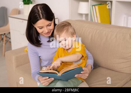 Joli petit garçon pointant sur l'image sur la page du livre tout en discutant de l'un des contes de fées avec sa mère pendant la période de l'isolement à la maison Banque D'Images