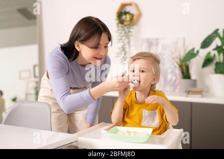 Jeune femme soigneuse en tenue de casual se courbant sur la petite table de dîner tout en aidant son petit-fils mignon avec le petit déjeuner dans la cuisine Banque D'Images