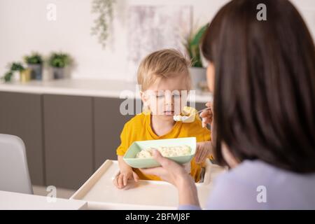 Mignon petit garçon mangeant de savoureux porridge maison tout en tenant la mère soigneuse cuillère par sa bouche tout en lui nourrissant pendant le petit déjeuner Banque D'Images