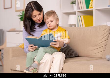 Jolie jeune mère lisant son petit livre de contes de fées tout en le tenant sur les genoux pendant le repos à la maison ou l'auto-isolation Banque D'Images