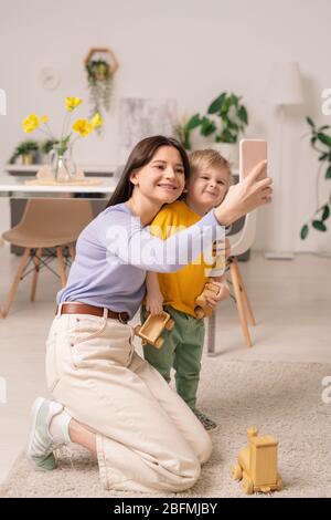 Jeune femme gaie et son adorable petit fils regardant l'appareil photo de smartphone tout en faisant du selfie chez soi pendant l'auto-isolation Banque D'Images