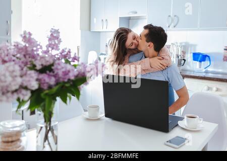Heureux couple de famille utilisant un ordinateur portable ayant du café dans la cuisine pendant la quarantaine du coronavirus. Jeune homme et femme embrassant Banque D'Images