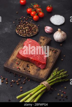 Tranche de steak de bœuf cru sur planche à découper en bois avec tomates, ail et pointes d'asperges sur fond sombre de table de cuisine. Banque D'Images