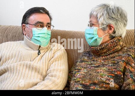 Covid-19, restez à la maison. Couple de personnes âgées joyeuses avec masques de protection, assis sur un canapé rire et se regardant, étant positif pendant la quarantaine à la maison . Banque D'Images