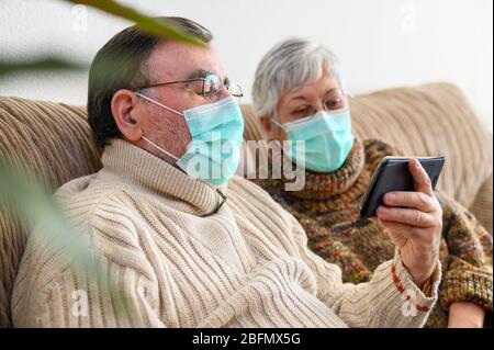 Coronavirus. Restez à la maison, style de vie.un couple de personnes âgées joyeux assis sur un canapé en quarantaine à la maison, en passant un appel vidéo avec le smartphone. Couple senior portant des masques de protection . Banque D'Images