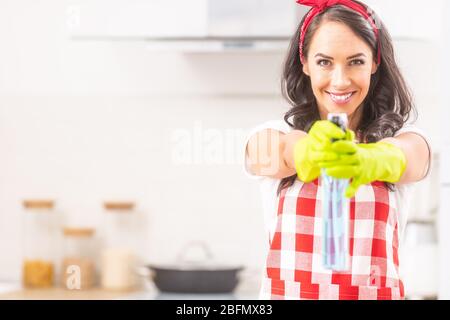 Femme de ménage souriante debout dans la cuisine pointant un détergent prêt à pulvériser des gants en caoutchouc vers l'appareil photo. Banque D'Images