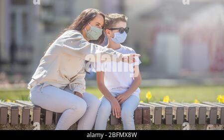 Mère et fils dans les masques de visage s'assoient sur un banc de ville avec elle pointant la direction qu'ils regardent tous les deux. Banque D'Images
