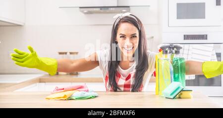 Une femme de ménage souriante qui tient ses bras bien ouverts dans la cuisine, avec des chiffons et des détergents colorés sur le bureau. Banque D'Images