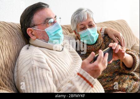 Coronavirus. Restez à la maison, style de vie.un couple de personnes âgées joyeux assis sur un canapé en quarantaine à la maison, en passant un appel vidéo avec le smartphone. Couple senior portant des masques de protection . Banque D'Images