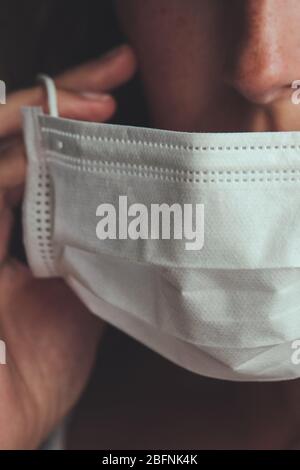 Photo détaillée d'une femme blanche non reconnaissable mettant sur un masque médical blanc pour empêcher la grippe. Fond flou, foyer sur le masque. Coronavirus, COVID-19 quarantaine. Concept de verrouillage. Banque D'Images