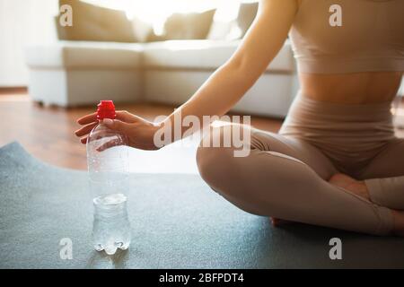 Jeune femme faisant de l'entraînement de yoga dans la salle pendant la quarantaine. Couper la vue de la fille dans le sport porter s'asseoir sur le tapis de yoga en position lotus et tenir la bouteille d'eau Banque D'Images