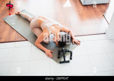 Jeune femme faisant de l'entraînement de yoga dans la salle pendant la quarantaine. Reposant sur un tapis avec une face vers le bas et maintenant une jambe sous le bras. Étirant une autre jambe dans le yoga Banque D'Images