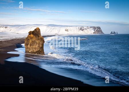 La mer pile et plage de sable noir avec des falaises enneigées derrière à Dyrhólaey en Islande Banque D'Images
