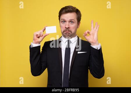 Homme d'âge mûr caucasien avec moustache drôle en costume montrant carte de visite donnant un geste parfait ou ok. Studio tourné Banque D'Images