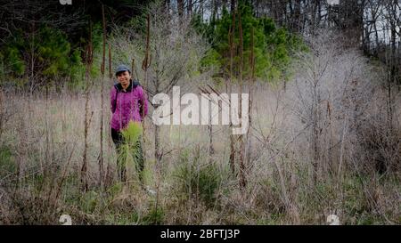 Femme en casquette et manteau violet debout au milieu du pinceau dans une forêt à côté d'un chemin dans le comté d'Orange, Caroline du Nord Banque D'Images