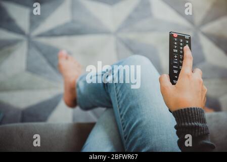 Vue de dessus de la jeune femme tenant la télécommande et allumant le téléviseur tout en étant assise sur un canapé chez elle. Arrêtez le virus Covid-19.Restez à la maison concept. Banque D'Images