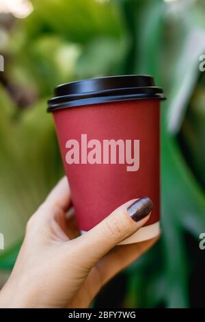 Main de la femme tenant une tasse de café rouge sur fond vert naturel. Banque D'Images