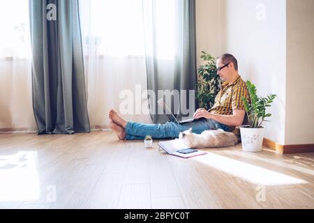 Le jeune homme siite sur le sol et travaille à la maison sur un ordinateur avec le téléphone, les papiers et une tasse de café. Travail à distance, travail à domicile. Banque D'Images