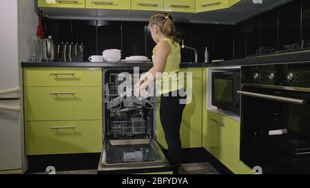 Une fille intelligente qui apprend à utiliser le lave-vaisselle. Appareils de cuisine modernes et élégants en noir vert. L'enfant met des plats propres. Banque D'Images