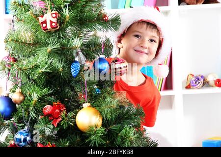 Un petit garçon à Santa Hat s'élève derrière l'arbre de Noël Banque D'Images