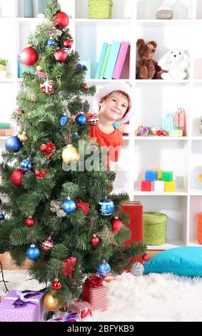 Un petit garçon à Santa Hat s'élève derrière l'arbre de Noël Banque D'Images