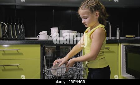 Une fille intelligente qui apprend à utiliser le lave-vaisselle. Appareils de cuisine modernes et élégants en noir vert. L'enfant met des plats sales. Banque D'Images