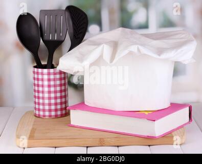 Chapeau de chef avec cuillères et livre de cuisine à bord sur table en bois sur fond de fenêtre Banque D'Images