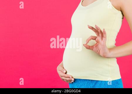 Image rognée d'une femme enceinte positive dans des vêtements colorés à la maison montrant un geste correct contre son ventre à fond rose. Grossesse facile et heureuse Banque D'Images