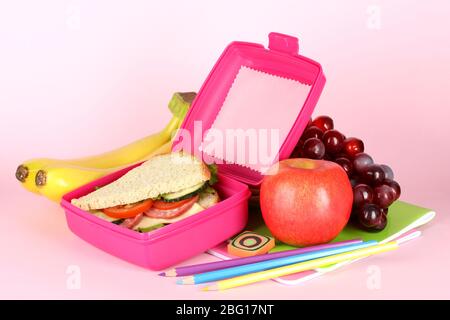 Boîte à déjeuner avec sandwich, fruits et articles de papeterie sur fond rose Banque D'Images