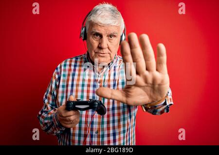 Joueur de haut niveau qui joue à un jeu vidéo à l'aide d'un joystick et d'un casque avec une main ouverte, faisant un signe d'arrêt avec une expression sérieuse et sûre, Banque D'Images