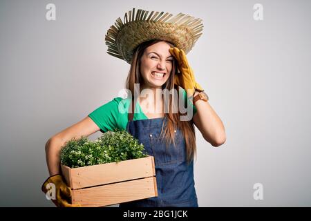 Jeune femme de campagne à tête rouge portant un tablier et une boîte de maintien de chapeau avec des plantes stressées avec la main sur la tête, choquée avec la honte et le visage surprise, un Banque D'Images