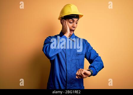 Jeune homme de travail africain américain portant un uniforme bleu et un casque de sécurité regardant l'heure de la montre inquiète, peur de se mettre en retard Banque D'Images