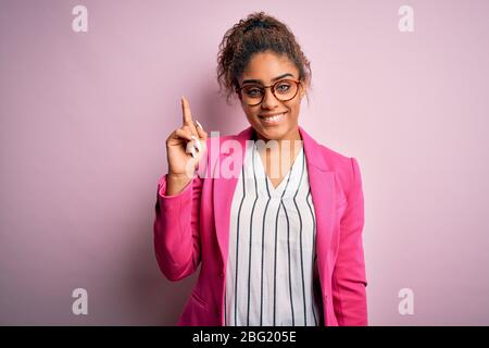 Belle femme d'affaires afro-américaine portant une veste et des lunettes sur fond rose montrant et pointant vers le haut avec le doigt numéro un tout en souriant Banque D'Images