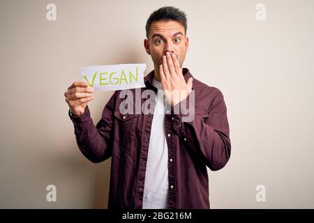 Jeune véggie homme tenant le papier avec le message vegan sur fond blanc couvrir la bouche avec la main choquée avec la honte pour l'erreur, l'expression de f Banque D'Images