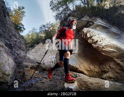 Vieil homme à barbe blanche en chemise rouge et sac à dos la traversée du fleuve dans la région de canyon. Outdoor travel concept Banque D'Images